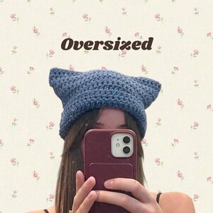 May include: A blue crochet cat ear hat with the word "Oversized" in brown text. The hat is made of a chunky yarn and has pointed ears. The background has a floral pattern.