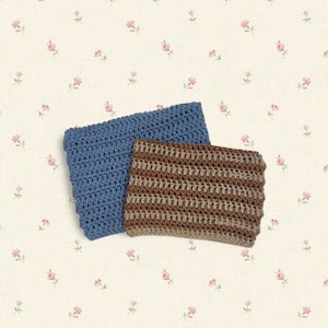 May include: Two crocheted cozies in the image. One is a solid blue, and the other is brown and tan striped. The cozies are rectangular and appear to be handmade. The background is a cream-colored fabric with small pink floral patterns.