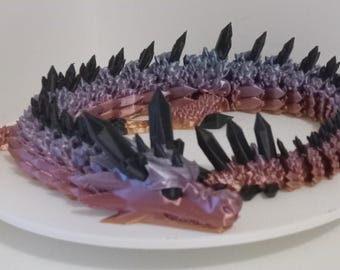 May include: A 3D printed dragon sculpture with a gradient of colors from black to purple to pink. The dragon is curled up in a circle and has a spiky back.