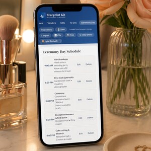 May include: A smartphone displays a wedding day schedule from the "Blueprint Kit" app. The screen shows a detailed timeline for the ceremony, including hair and makeup, first look, ceremony, and reception details. The phone is on a marble surface with flowers.