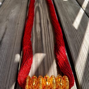 May include: A long red beaded necklace with a gold metal clasp and a decorative gold metal pendant. The pendant is made up of several round pieces with intricate designs.