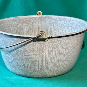 May include: A vintage, speckled gray enamelware pot with a wire handle and two side handles. The pot has a rounded shape and a textured surface. The handles are black and white. The pot is set against a green background.