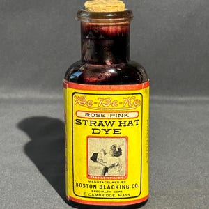 Puede incluir: Una botella de vidrio vintage de tinte para sombreros de paja rosa Be-Be-Ko. La botella tiene un tapón de corcho y una etiqueta amarilla con texto negro y rojo. La etiqueta incluye una ilustración y el nombre del fabricante, Boston Blacking Co.
