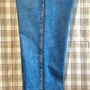 K&ouml;nnte beinhalten: Eine blaue Jeanshose mit einer Ges&auml;&szlig;tasche. Die Jeans h&auml;ngt an einem Kleiderb&uuml;gel vor einem karierten Hintergrund.
