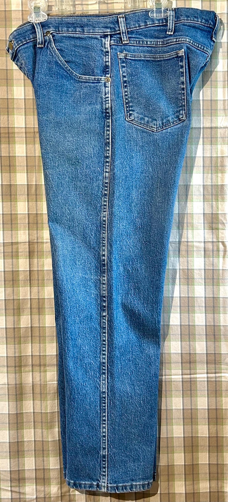 K&ouml;nnte beinhalten: Eine blaue Jeanshose mit einer Ges&auml;&szlig;tasche. Die Jeans h&auml;ngt an einem Kleiderb&uuml;gel vor einem karierten Hintergrund.