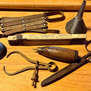 May include: A collection of vintage tools including a metal accordion-style gate, a small funnel, a Lufkin Rule Co. No. 524 folding wooden ruler, a metal oil can, a wooden-handled awl, and a pair of metal scissors with a brass caliper tool.