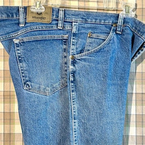 K&ouml;nnte beinhalten: Eine blaue Jeanshose mit einem braunen Leder-Patch auf der Ges&auml;&szlig;tasche, auf dem "Wrangler" steht.