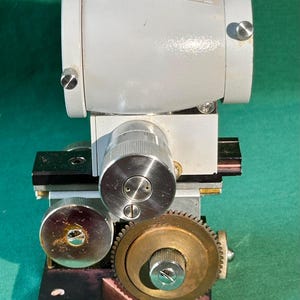 May include: A vintage, industrial-style optical instrument with a white cylindrical body and multiple silver knobs and gears. The device is mounted on a dark base and features a brass gear. The background is a solid green.