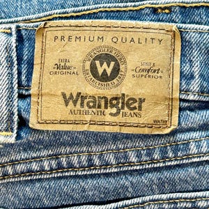 K&ouml;nnte beinhalten: Ein braunes Leder-Etikett mit dem Text "PREMIUM QUALITY" und "Wrangler AUTHENTIC JEANS" in Wei&szlig;. Das Etikett zeigt auch ein schwarz-wei&szlig;es Logo mit dem Text "Wrangler Hero Established 1947" und dem Text "Extra Value Original" und "Style & Comfort Superior".