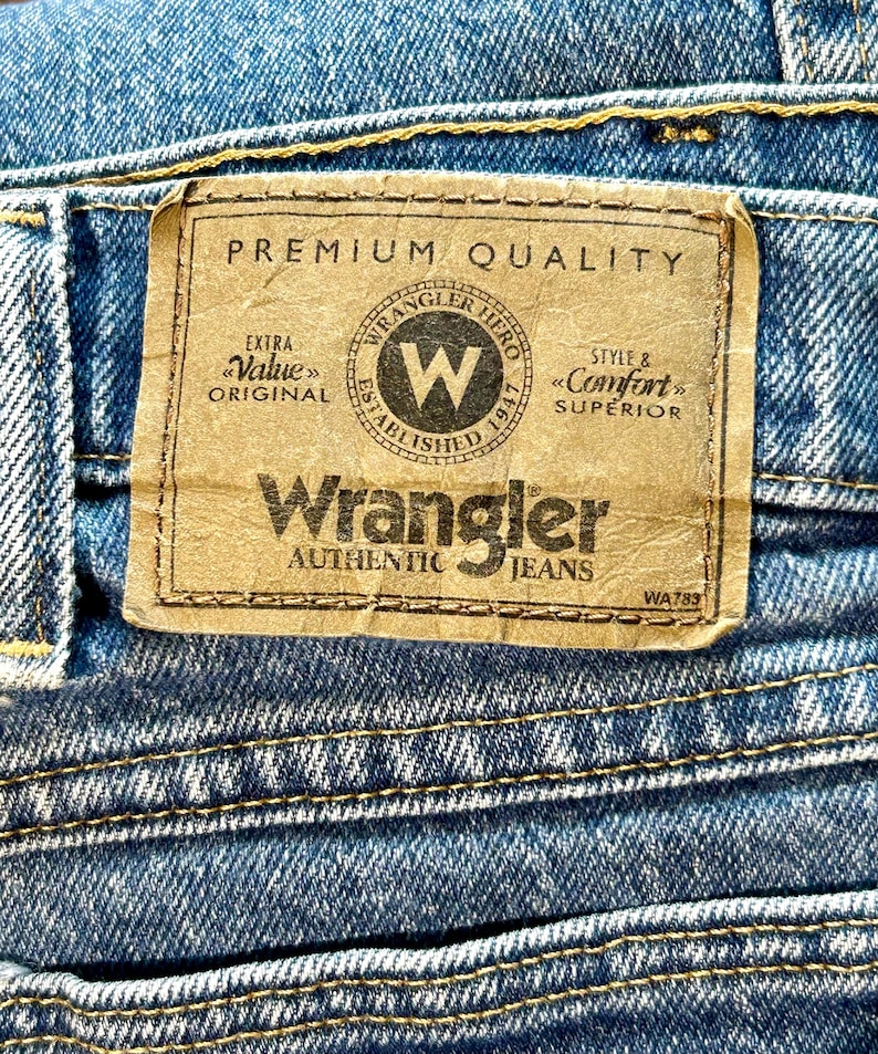 K&ouml;nnte beinhalten: Ein braunes Leder-Etikett mit dem Text "PREMIUM QUALITY" und "Wrangler AUTHENTIC JEANS" in Wei&szlig;. Das Etikett zeigt auch ein schwarz-wei&szlig;es Logo mit dem Text "Wrangler Hero Established 1947" und dem Text "Extra Value Original" und "Style & Comfort Superior".