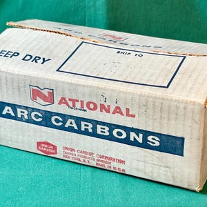 Op de afbeelding: Een rechthoekige kartonnen doos met de tekst "KEEP DRY" en "SHIP TO" gedrukt op de bovenkant. De doos heeft het "NATIONAL ARC CARBONS" logo in rood en blauw. De doos staat op een groene ondergrond.