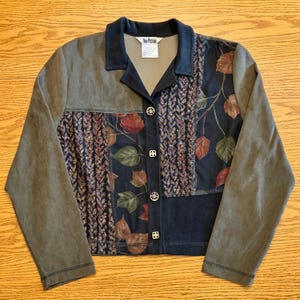 May include: A long-sleeve jacket featuring a dark blue collar, olive green sleeves, and a front panel with a mix of dark blue fabric with a leaf print and textured vertical strips. The jacket has four silver-toned buttons and a Nina Piccino label.