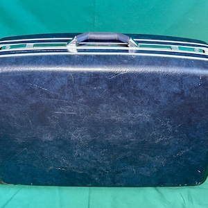 May include: A vintage navy blue suitcase with white and silver accents. The suitcase has a handle on top and is rectangular in shape. The suitcase is sitting on a green surface.
