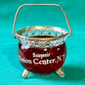 May include: A small red glass cauldron with a gold rim and a wire handle. The cauldron has three gold feet and says "Souvenir Union Center, N.Y." on the side.