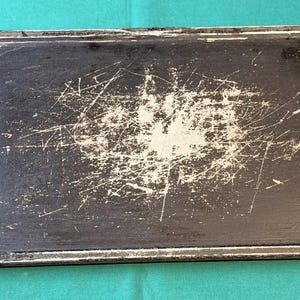 Puede incluir: Una bandeja de horno rectangular de metal negro con un borde elevado. La superficie muestra un desgaste significativo, con numerosas rayas blancas y áreas donde el acabado oscuro se ha desgastado, concentradas en el centro de la bandeja.
