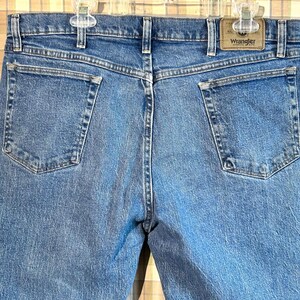 K&ouml;nnte beinhalten: Eine blaue Jeanshose mit einem braunen Leder-Patch auf der rechten Ges&auml;&szlig;tasche, auf dem "Wrangler" steht.