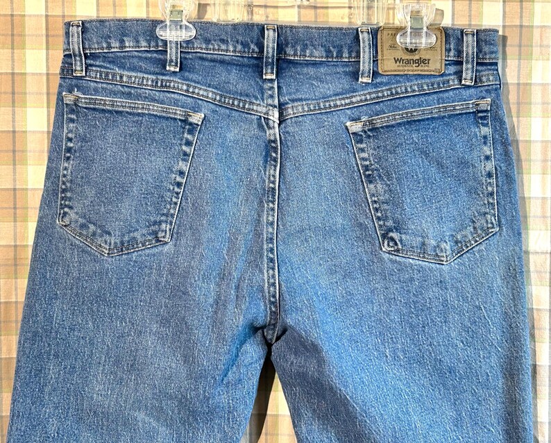 K&ouml;nnte beinhalten: Eine blaue Jeanshose mit einem braunen Leder-Patch auf der rechten Ges&auml;&szlig;tasche, auf dem "Wrangler" steht.