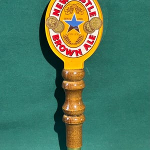 Puede incluir: Un tirador de cerveza Newcastle Brown Ale. La parte superior ovalada amarilla presenta el logotipo de la marca con una estrella azul y las palabras "Newcastle Brown Ale". El mango es de madera con una punta de metal color latón.