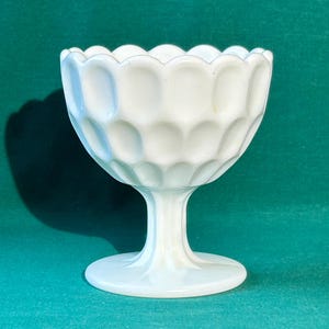 May include: A white, vintage-style glass goblet with a scalloped rim and a textured, honeycomb-like pattern. The goblet has a pedestal base and a stem. The glass is opaque and appears to be milk glass.