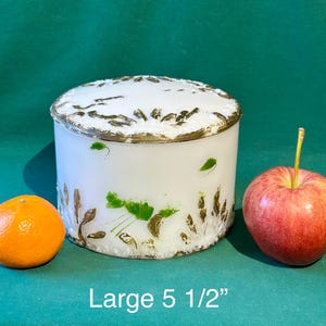 May include: White ceramic jar with gold trim and floral design. The jar is 5 1/2 inches in diameter.  The jar is sitting on a green surface with a red apple and an orange.