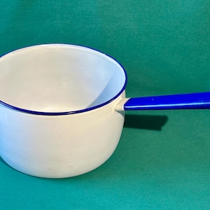 May include: White enamel saucepan with a blue handle and rim. The pot has a rounded shape and a long, straight handle. The handle has a hole at the end. The saucepan is on a green surface.