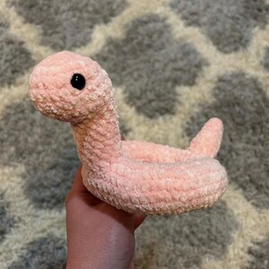 May include: A handmade, plush pink worm toy with a black eye. The worm is made of a soft, textured material and has a curved body with a small tail. The toy is designed to be cuddly and is suitable for children.