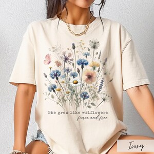 Puede incluir: Camiseta de color marfil con un dise&ntilde;o floral en acuarela con flores azules, rosas y amarillas. El texto "She grew like wildflowers, fierce and free" est&aacute; impreso debajo. La camiseta tiene mangas cortas.