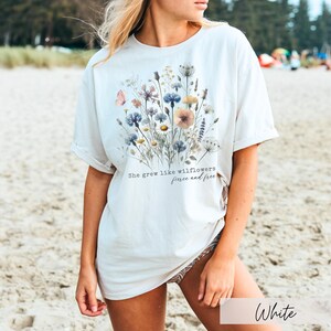 Puede incluir: Camiseta blanca con un dise&ntilde;o floral y el texto "She grew like wildflowers, fierce and free." El dise&ntilde;o incluye flores azules, amarillas y rosas. La camiseta est&aacute; hecha de un material suave.