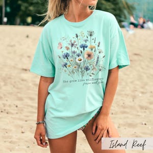 Puede incluir: Camiseta turquesa claro con dise&ntilde;o floral y el texto "She grew like wildflowers fierce and free". La camiseta tiene mangas cortas y un corte relajado, ideal para uso diario.