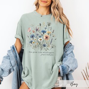 Puede incluir: Camiseta verde salvia con un dise&ntilde;o floral en acuarela con flores rosas, azules y amarillas. El texto "She grew like wildflowers, fierce and free" est&aacute; impreso debajo. La camiseta tiene un corte holgado.