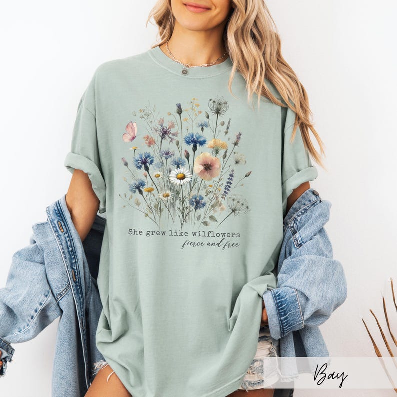 Puede incluir: Camiseta verde salvia con un dise&ntilde;o floral en acuarela con flores rosas, azules y amarillas. El texto "She grew like wildflowers, fierce and free" est&aacute; impreso debajo. La camiseta tiene un corte holgado.