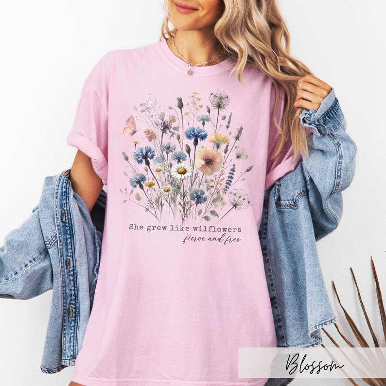 Puede incluir: Camiseta rosa claro con un dise&ntilde;o floral de flores silvestres azules, amarillas y blancas. El texto de la camiseta dice: "She grew like wildflowers, fierce and free."