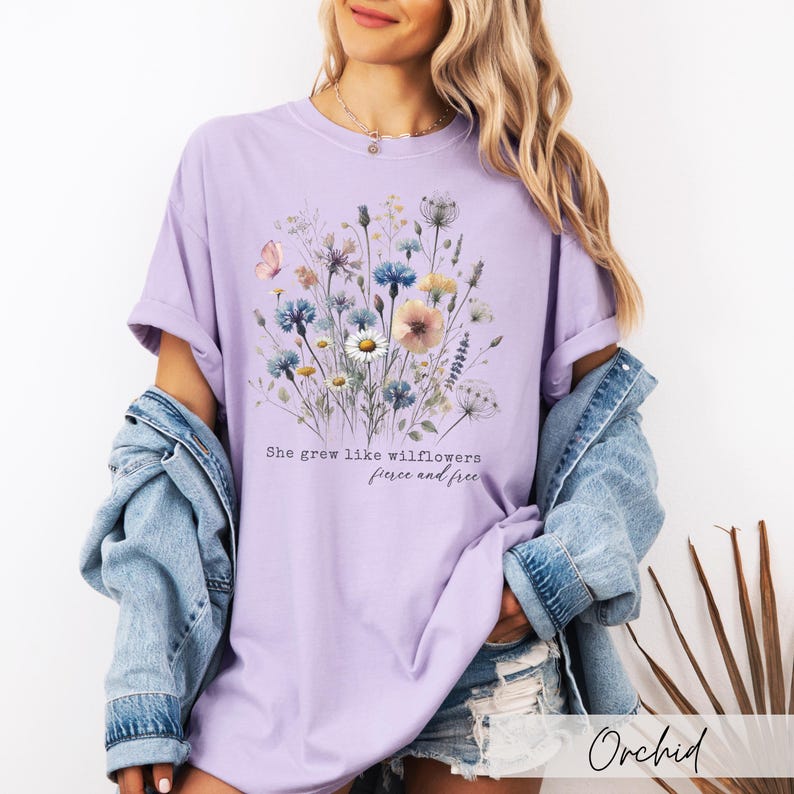 Puede incluir: Camiseta color orqu&iacute;dea con un dise&ntilde;o floral y una mariposa. El dise&ntilde;o incluye varias flores silvestres en tonos azules, rosas y blancos. El texto "She grew like wildflowers, fierce and free" est&aacute; impreso debajo.