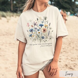 Puede incluir: Camiseta color marfil con un dise&ntilde;o floral en acuarela con flores azules, rosas y amarillas. El texto "She grew like wildflowers, fierce and free" est&aacute; impreso debajo. La camiseta tiene mangas cortas.