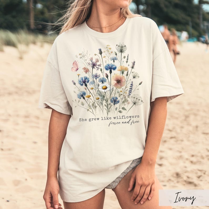 Puede incluir: Camiseta color marfil con un dise&ntilde;o floral en acuarela con flores azules, rosas y amarillas. El texto "She grew like wildflowers, fierce and free" est&aacute; impreso debajo. La camiseta tiene mangas cortas.