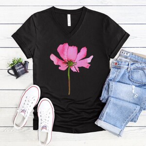 May include: Black v-neck t-shirt with a pink watercolor flower graphic. The shirt is styled with blue jeans and white sneakers.