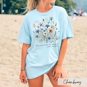 Puede incluir: Camiseta azul claro con dise&ntilde;o floral y el texto "She grew like wildflowers, fierce and free". El gr&aacute;fico presenta varias flores en tonos azules, rosas y amarillos. La camiseta es de corte holgado.