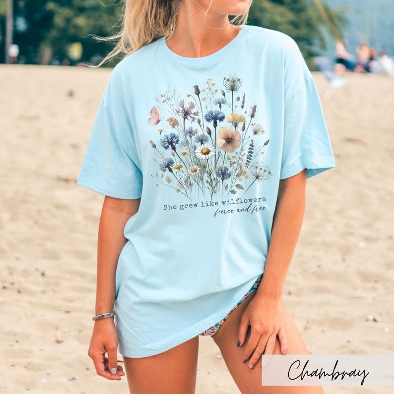 Puede incluir: Camiseta azul claro con dise&ntilde;o floral y el texto "She grew like wildflowers, fierce and free". El gr&aacute;fico presenta varias flores en tonos azules, rosas y amarillos. La camiseta es de corte holgado.