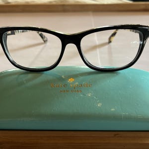 Może przedstawiać: Czarne okulary w kształcie kociego oka spoczywają na turkusowym etui na okulary Kate Spade. Okulary mają wzór na zausznikach i przezroczyste soczewki. Etui ma złote logo Kate Spade.