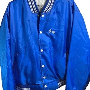 May include: A vibrant blue satin bomber jacket with white and blue striped trim on the collar, cuffs, and hem. The jacket features white snap closures and the name "Jay" embroidered in white script on the chest. The jacket has a classic varsity style.