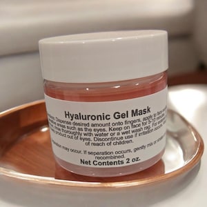 May include: A jar of Hyaluronic Gel Mask with a white lid and a pinkish gel inside. The label reads "Hyaluronic Gel Mask" and "Net Contents 2 oz.". The jar sits on a copper-colored tray.