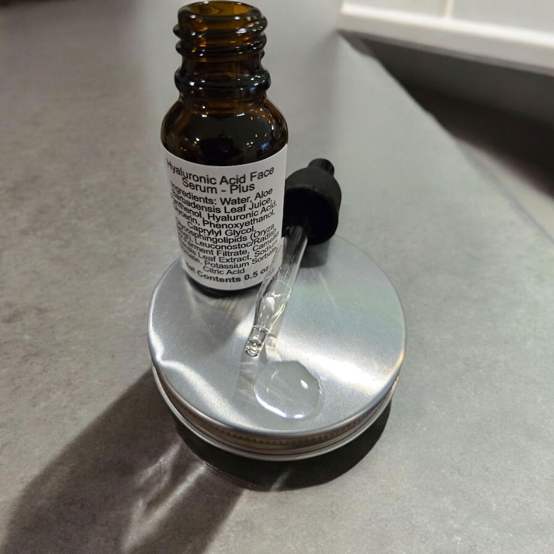 Peut inclure: Un flacon en verre brun avec un compte-gouttes, &eacute;tiquet&eacute; "Hyaluronic Acid Face Serum - Plus". Le flacon est pos&eacute; sur un couvercle argent&eacute; avec une &eacute;tiquette blanche.