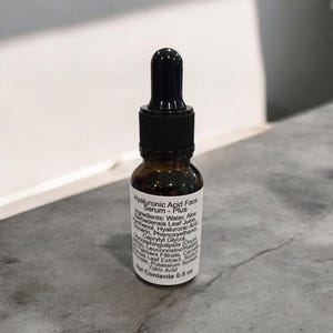 Peut inclure: Un petit flacon en verre brun avec un compte-gouttes noir. Le flacon est &eacute;tiquet&eacute; "Hyaluronic Acid Face Serum - Plus" et liste les ingr&eacute;dients. Le flacon contient 0,5 oz de produit.