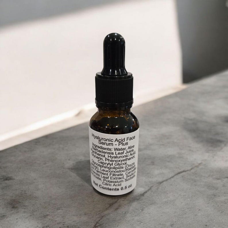 Peut inclure: Un petit flacon en verre brun avec un compte-gouttes noir. Le flacon est &eacute;tiquet&eacute; "Hyaluronic Acid Face Serum - Plus" et liste les ingr&eacute;dients. Le flacon contient 0,5 oz de produit.