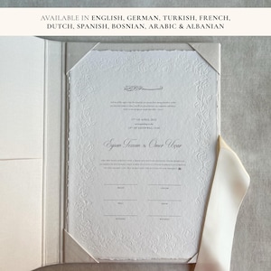 May include: A light grey wedding invitation with embossed floral detailing. The invitation is open, revealing printed text and a cream-coloured ribbon. The text includes names and dates. The top of the invitation reads "AVAILABLE IN ENGLISH, GERMAN, TURKISH, FRENCH, DUTCH, SPANISH, BOSNIAN, ARABIC & ALBANIAN."