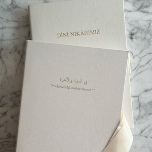 Peut inclure: Deux livres de mariage blanc cassé avec des lettres dorées. Le livre du haut indique "DİNİ NİKAHIMIZ". Le livre du bas contient du texte en arabe et en anglais : "in this world, and in the next."