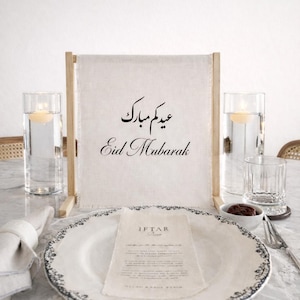 Peut inclure: Une table dressée avec une assiette blanche bordée de motifs floraux noirs, une serviette en lin et un menu. Un cadre en bois maintient une bannière en lin avec "Eid Mubarak" en arabe et en anglais. Deux vases en verre avec des bougies flottantes.