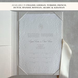 Könnte beinhalten: Eine weiße Einladung mit geprägten floralen Details und Text in mehreren Sprachen. Die Einladung befindet sich in einem weißen Ordner und ist mit einem weißen Band gebunden. Der Text enthält Namen und Veranstaltungsdetails.