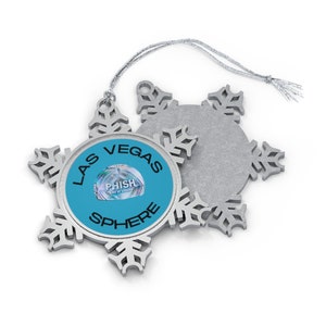 May include: Silver snowflake ornament with a blue circle in the center that says "LAS VEGAS SPHERE" with a smaller image of a concert poster for Phish at the Sphere.
