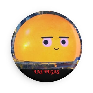 May include: A round button featuring a cartoon character with a yellow, dome-shaped head, black eyebrows, and pink eyes. The word "LAS VEGAS" is printed in red at the bottom, set against a black background. The background shows a cityscape.